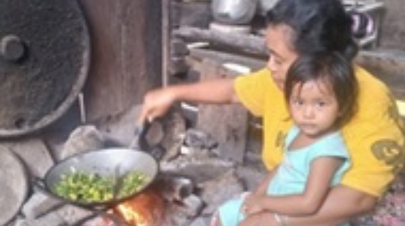 mama-Nela-1-1@3x Seorang wanita sedang memasak sambil menggendong seorang anak.